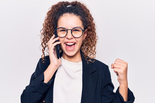 Beautiful kid girl with curly hair wearing glasses having conversation talking on the smartphone screaming proud, celebrating victory and success very excited with raised arms