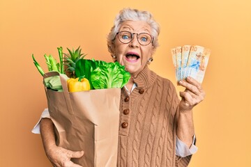 Senior grey-haired woman holding groceries and swiss franc banknotes celebrating crazy and amazed...