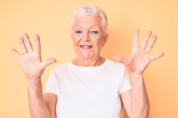 Senior beautiful woman with blue eyes and grey hair wearing classic white tshirt over yellow background showing and pointing up with fingers number ten while smiling confident and happy.