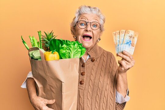 Senior grey-haired woman holding groceries and swiss franc banknotes celebrating crazy and amazed for success with open eyes screaming excited.