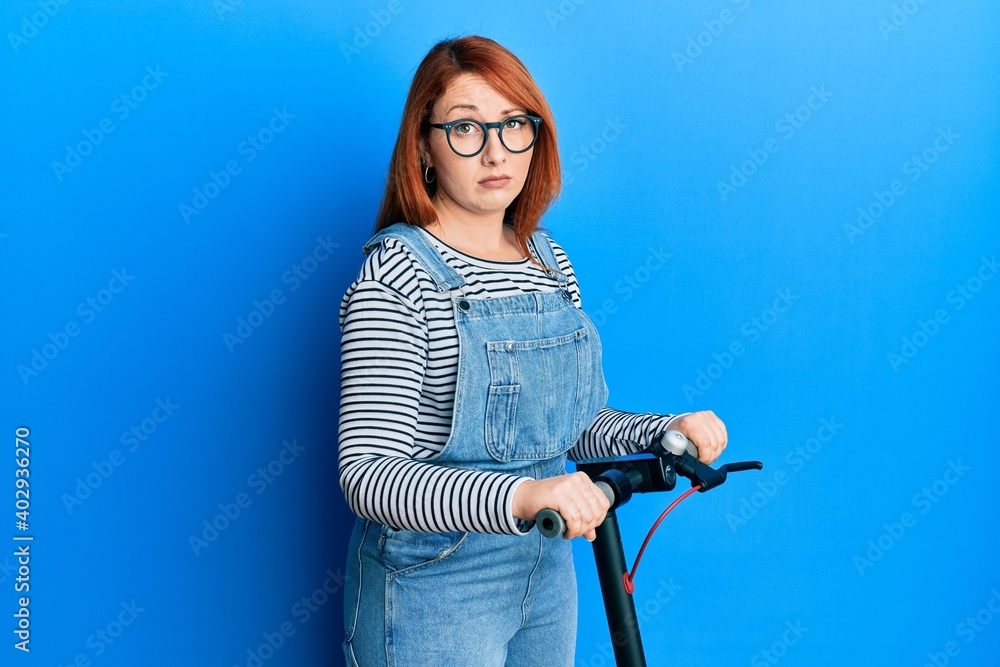 Sticker beautiful redhead woman standing on modern scooter over blue background skeptic and nervous, frownin