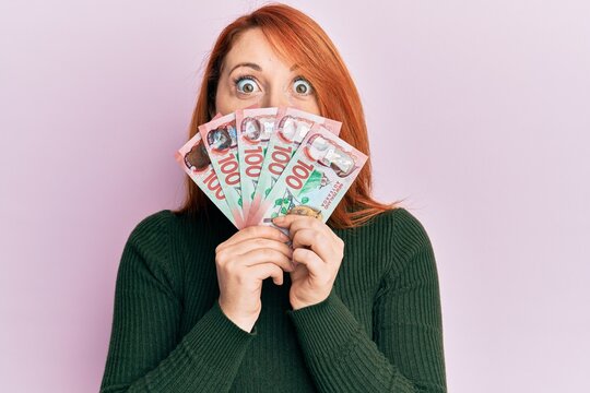 Beautiful redhead woman holding 100 new zealand dollars banknote afraid and shocked with surprise and amazed expression, fear and excited face.