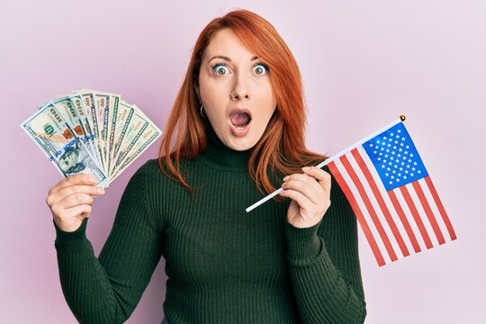 Beautiful redhead woman holding dollars banknote and usa flag afraid and shocked with surprise and amazed expression, fear and excited face.