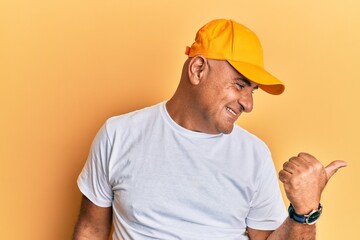 Mature middle east man with mustache wearing casual white tshirt and yellow cap pointing thumb up to the side smiling happy with open mouth