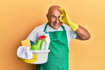 Mature middle east man wearing cleaner apron holding cleaning products smiling happy doing ok sign with hand on eye looking through fingers