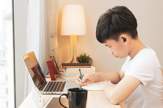 A Handsome Asian Teenager Boy With Wireless Earbuds Concentrate Study From Home With Virtual Classroom Through Computer Laptop. Covid 19 Pandemic Lockdown. New Normal Lifestyle, Social Distancing.