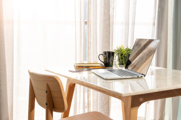 Beautiful workspace - online remote work from home concept. A wooden table with computer laptop, smartphone, coffee cup beside the window balcony with sun light shine through the curtain. No people.