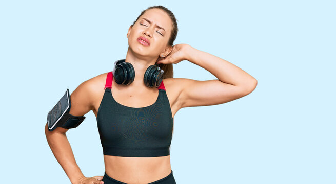 Beautiful blonde woman wearing gym clothes and using headphones suffering of neck ache injury, touching neck with hand, muscular pain