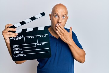 Middle age bald man holding video film clapboard covering mouth with hand, shocked and afraid for...