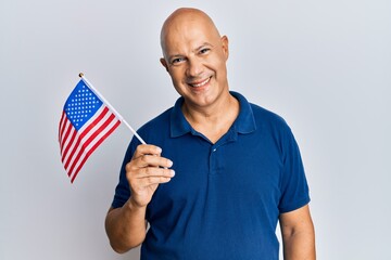 Middle age bald man holding united states flag looking positive and happy standing and smiling with a confident smile showing teeth