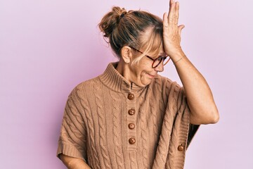 Middle age blonde woman wearing glasses over pink background surprised with hand on head for...
