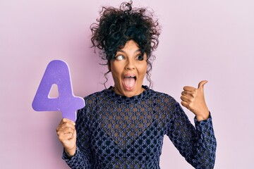 Young hispanic woman holding number four pointing thumb up to the side smiling happy with open mouth