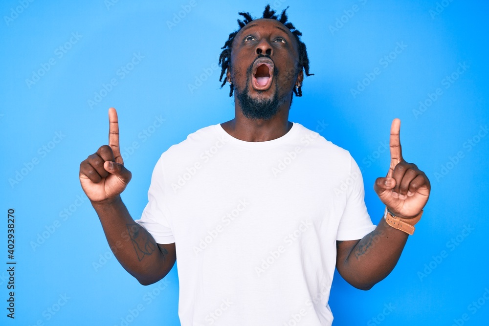 Poster Young african american man with braids wearing casual white tshirt amazed and surprised looking up and pointing with fingers and raised arms.