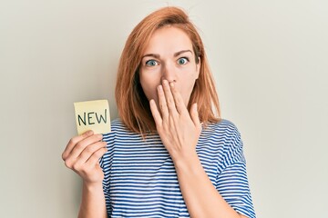Young caucasian woman holding reminder with new word covering mouth with hand, shocked and afraid...