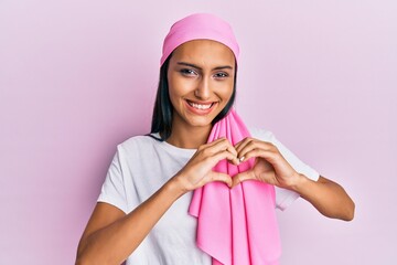 Young brunette woman wearing breast cancer support pink scarf smiling in love showing heart symbol and shape with hands. romantic concept.