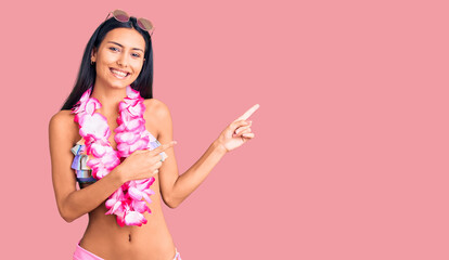 Young beautiful latin girl wearing bikini and hawaiian lei smiling and looking at the camera pointing with two hands and fingers to the side.