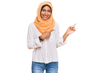 Young brunette woman wearing traditional islamic hijab scarf smiling and looking at the camera pointing with two hands and fingers to the side.