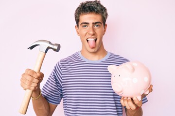 Young handsome man holding piggy bank and hammer sticking tongue out happy with funny expression.