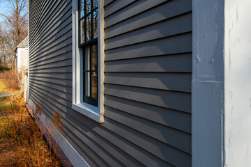 Corner view of a vintage tan coloured house with white trim and black wooden window spacers. The glass in the window is wavy and reflecting the sun. There are orange and red leaves on the ground.