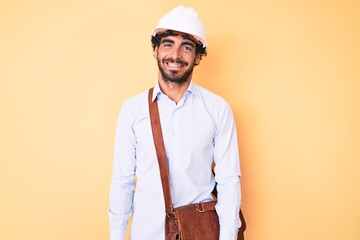 Handsome young man with curly hair and bear wearing architect hardhat looking positive and happy standing and smiling with a confident smile showing teeth