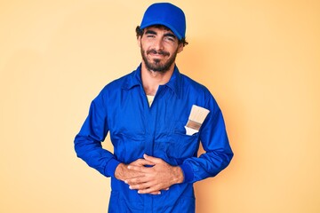 Handsome young man with curly hair and bear wearing builder jumpsuit uniform with hand on stomach because indigestion, painful illness feeling unwell. ache concept.