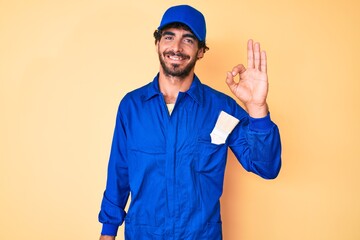 Handsome young man with curly hair and bear wearing builder jumpsuit uniform smiling positive doing ok sign with hand and fingers. successful expression.