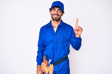 Handsome young man with curly hair and bear weaing handyman uniform showing and pointing up with finger number one while smiling confident and happy.
