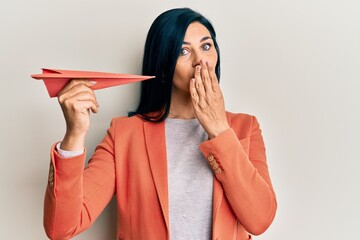 Young caucasian woman holding paper airplane covering mouth with hand, shocked and afraid for...