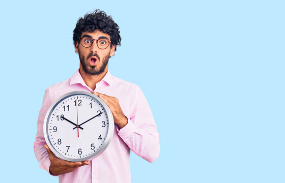 Handsome young man with curly hair and bear holding big clock scared and amazed with open mouth for surprise, disbelief face