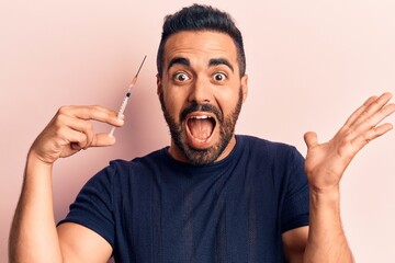 Young hispanic man holding syringe celebrating victory with happy smile and winner expression with raised hands