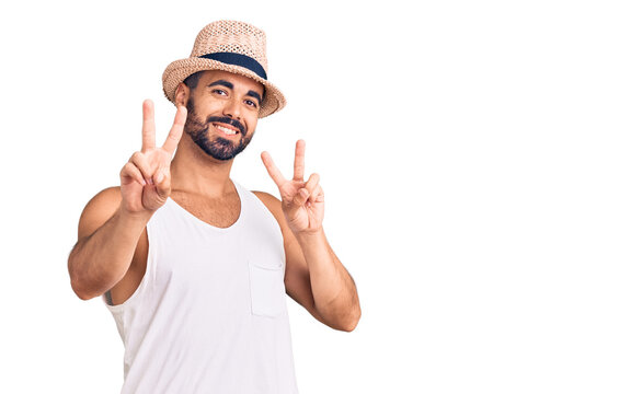 Young hispanic man wearing casual summer hat smiling looking to the camera showing fingers doing victory sign. number two.