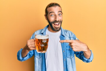 Attractive man with long hair and beard drinking a pint of beer smiling happy pointing with hand and finger