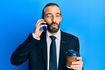 Naklejka premium Attractive business man with long hair and beard using smartphone and drinking a cup of coffee looking at the camera blowing a kiss being lovely and sexy. love expression.