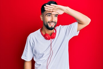 Young man with beard listening to music using headphones very happy and smiling looking far away with hand over head. searching concept.