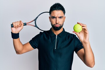 Young man with beard playing tennis holding racket and ball puffing cheeks with funny face. mouth inflated with air, catching air.