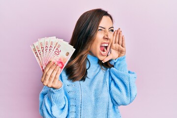 Young brunette woman holding 20 israel shekels banknotes shouting and screaming loud to side with hand on mouth. communication concept.