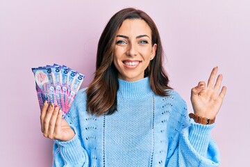 Young brunette woman holding hong kong 10 dollars banknotes doing ok sign with fingers, smiling friendly gesturing excellent symbol