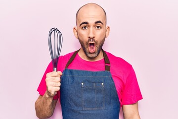 Young handsome bald man wearing apron holding whisk scared and amazed with open mouth for surprise, disbelief face