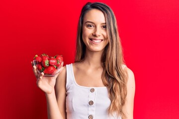 Young beautiful girl holding bowl with strawberries looking positive and happy standing and smiling...