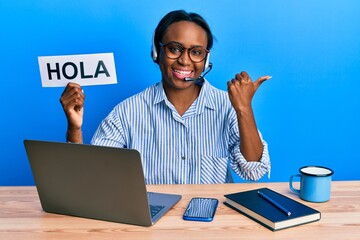 Young african woman wearing operator headset holding hola word pointing thumb up to the side smiling happy with open mouth © Krakenimages.com