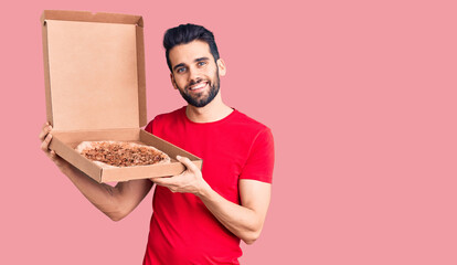 Young handsome man with beard holding delivery cardoboard with italian pizza looking positive and happy standing and smiling with a confident smile showing teeth