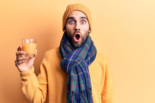 Young handsome man with beard wearing scarf and wool cap drinking orange juice scared and amazed with open mouth for surprise, disbelief face