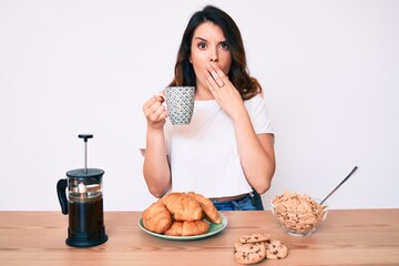 Young beautiful brunette woman sitting on the table having breakfast covering mouth with hand,...