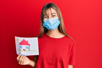 Teenager caucasian girl wearing medical mask and holding house draw looking positive and happy standing and smiling with a confident smile showing teeth