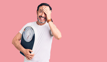 Middle age handsome man holding weighing machine stressed and frustrated with hand on head,...