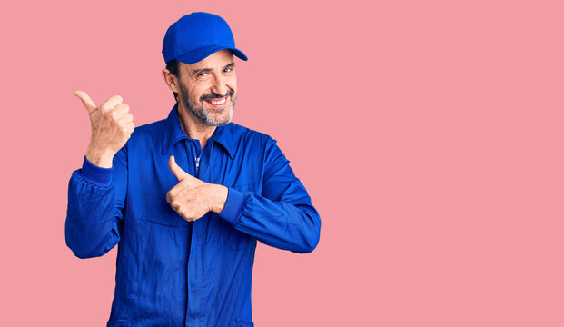 Middle age handsome man wearing mechanic uniform pointing to the back behind with hand and thumbs up, smiling confident
