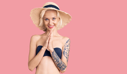 Young blonde woman with tattoo wearing bikini and summer hat praying with hands together asking for forgiveness smiling confident.