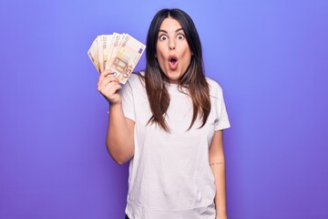 Young beautiful brunette woman holding euros banknotes over isolated purple background scared and amazed with open mouth for surprise, disbelief face