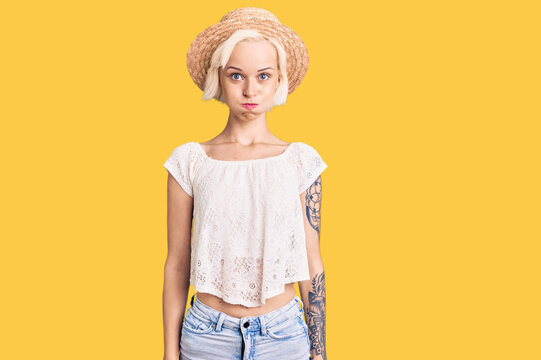 Young blonde woman with tattoo wearing summer hat puffing cheeks with funny face. mouth inflated with air, crazy expression.
