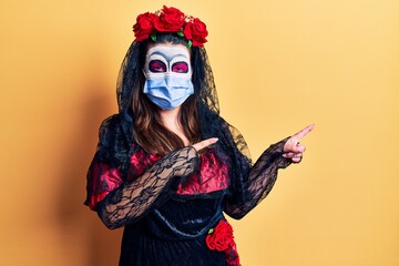 Young woman wearing day of the dead costume wearing medical mask smiling and looking at the camera pointing with two hands and fingers to the side.